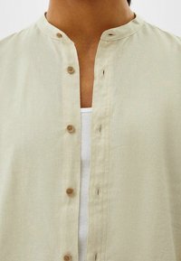 Lichtbeige overhemd met knopen en een mandarijnkraag, voorzien van houten knopen en een gestructureerde stof. Draagbaar over een witte geribbelde tanktop.