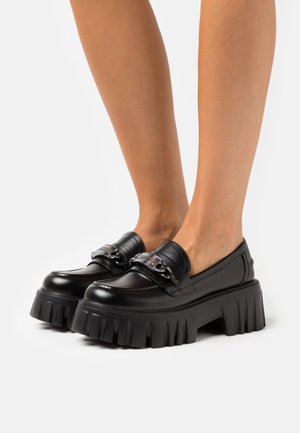 Mocassins en cuir noir avec une semelle épaisse et striée et un détail de chaîne métallique sur le dessus, présentant une texture lisse et un bout arrondi.