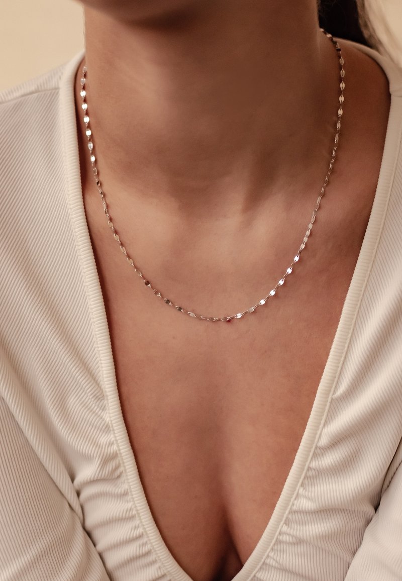 Collier en argent composé de maillons ovales et en forme de cœur, réfléchissant la lumière. Porté avec un haut blanc texturé à décolleté en V profond.