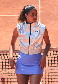 Silberner, ärmelloser, mit Reißverschluss versehenen Sportweste mit geripptem Saum, kombiniert mit einem hellblauen gerippten Rock. Der Hintergrund zeigt einen Tennisplatz.