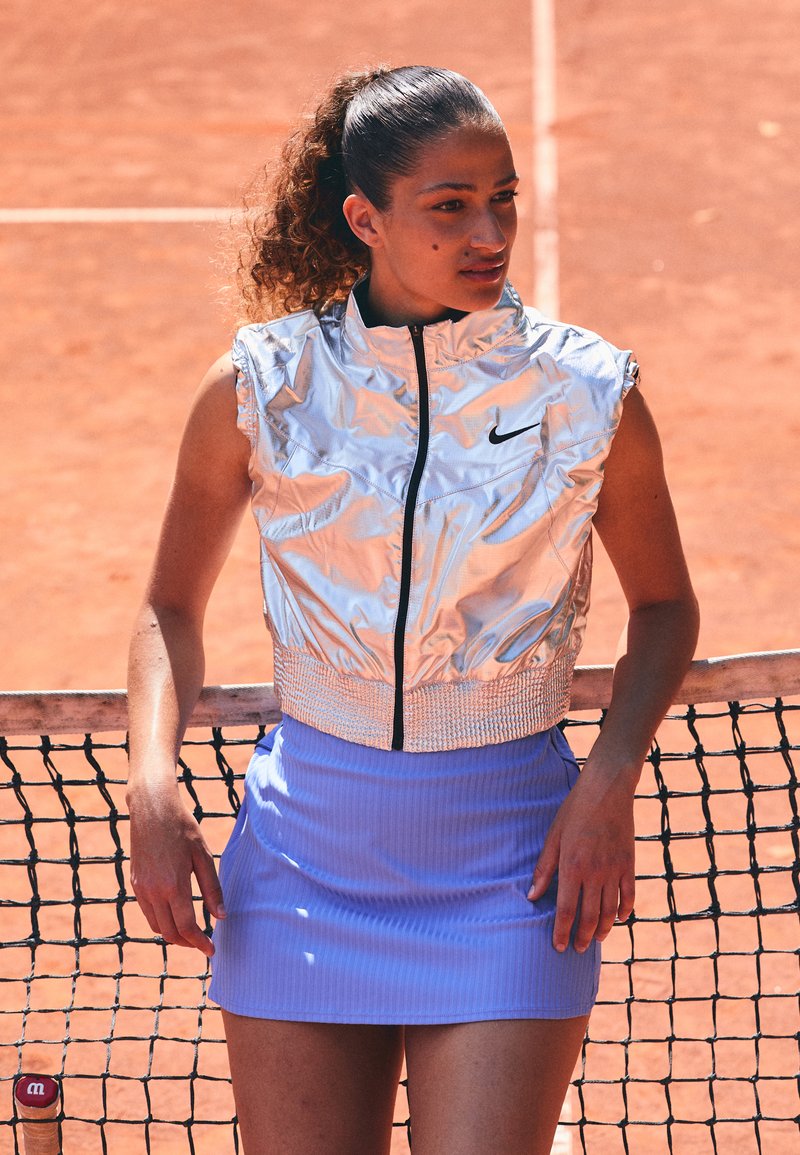Silberner, ärmelloser, mit Reißverschluss versehenen Sportweste mit geripptem Saum, kombiniert mit einem hellblauen gerippten Rock. Der Hintergrund zeigt einen Tennisplatz.