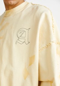 Sudadera color crema con un gráfico de la letra "A", bordes deshilachados y patrones de hojas sutiles. Fabricada con un material suave y texturizado.