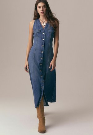 Femeie purtând o rochie albastră din denim fără mâneci, cu nasturi, și cizme de piele întoarsă maro până la genunchi, stând în picioare pe un fundal simplu.