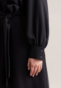 Schwarzes Kleid mit langen, gepufften Ärmeln und einem Taillengürtel. Die Manschette hat zwei Knöpfe und einen glatten, fließenden Stoff. 