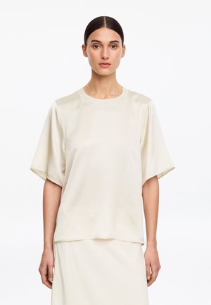 Femme aux cheveux foncés tirés en arrière portant un haut en satin ivoire à manches courtes et une jupe assortie, regardant devant avec une expression neutre.