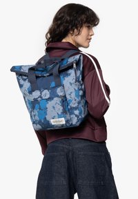 Mochila tipo roll-top en estampado de camuflaje azul, con un tejido resistente, dos asas superiores, correas ajustables y un bolsillo con cremallera en la parte frontal.