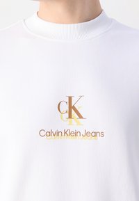 Chemise blanche à col montant avec le logo marron et jaune « cK Calvin Klein Jeans » centré sur la poitrine.