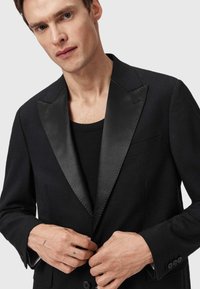 Uomo che allaccia il bottone di un blazer nero con risvolti in satin, indossando una camicia nera sotto, su uno sfondo neutro.
