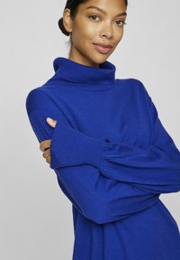 Kungligt blått polo-tröja med ribbade muddar och löst sittande ärmar. Mjuk textur; avslappnad design som erbjuder komfort och värme.
