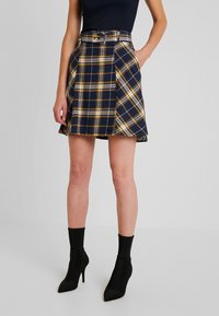 Gonna skater a quadri navy con linee gialle e bianche, design plissettato, cintura in vita, tasche laterali, abbinata a stivali neri alla caviglia.