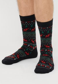 Fekete zoknik ünnepi mintával, melyen vörös rénszarvasok, zöld fák és fehér hópehely látható, textúrált kötött anyaggal és kontrasztos szegéllyel.