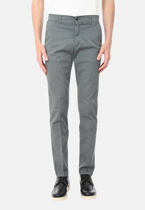 Uomo che indossa pantaloni grigi slim fit e scarpe nere stringate, in piedi con le braccia rilassate lungo i fianchi davanti a uno sfondo semplice.