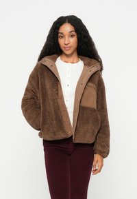 Veste en polaire marron avec un panneau avant imitation suède beige, boutons à pression, capuche et poches latérales, portée sur un haut blanc et un pantalon bordeaux.