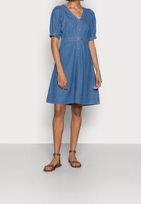 Denim-mekko, jossa on lyhyet puffigönit, V-pääntie ja napit edessä. A-linjainen malli keskisinisessä, joka on yhdistetty ruskeisiin latteisiin sandaaleihin.