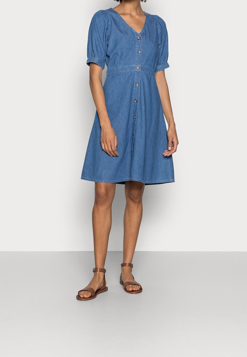 Denim-mekko, jossa on lyhyet puffigönit, V-pääntie ja napit edessä. A-linjainen malli keskisinisessä, joka on yhdistetty ruskeisiin latteisiin sandaaleihin.