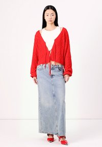 Cardigã cropped tricotado vermelho com um laço à frente, combinado com uma saia longa de denim. A modelo usa sapatos de salto agudo vermelhos, exibindo uma paleta de cores minimalista.