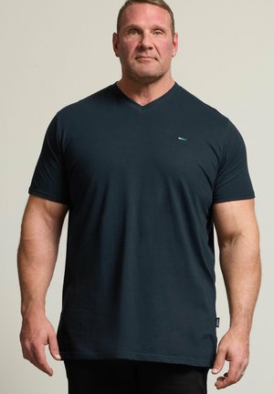 Muskulöser Mann mit kurzen Haaren steht frontal, trägt ein dunkelblaues T-Shirt mit kurzen Ärmeln und V-Ausschnitt vor einem schlichten Hintergrund.