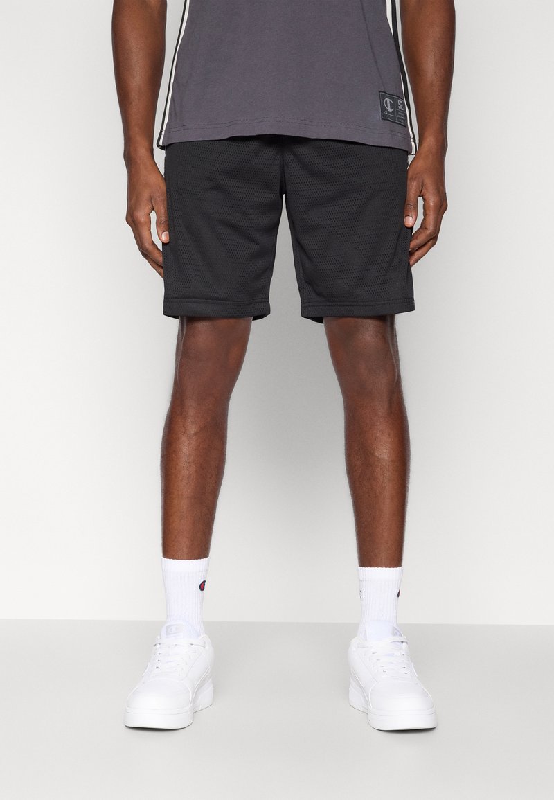 Μαύροι αθλητικοί σορτς από δίχτυ με ελαστική ζώνη μέσης, συνδυασμένοι με γκρι κοντομάνικο μπλουζάκι και λευκά αθλητικά π απούτσια. Λευκές κάλτσες με ορατό λογότυπο.