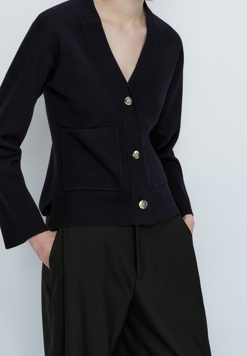 Navy knitted cardigan with a deep V-neck, three gold buttons, and two front pockets. Long sleeves with ribbed cuffs and hem.