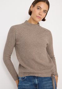 Brązowy sweter z prążkowanego materiału, z wysokim kołnierzem, bocznym zamkiem błyskawicznym i ściągaczem u dołu. Połączony z niebieskimi dżinsami. Miękki, teksturowany materiał.