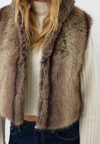 Brown faux fur vest with a cropped design. Features a soft, fluffy texture and wide collar. Worn over a ribbed white sweater and blue jeans.