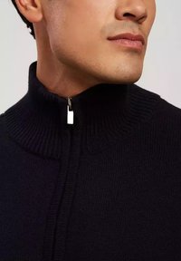 Granatowy sweter dzianinowy z wysokim kołnierzem i zamkiem błyskawicznym z przodu, z akcentem w postaci srebrnego wykończenia. Gładka faktura, dopasowany krój w okolicy szyi.