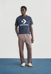 Μπλε βαμβακερό t-shirt με λευκό λογότυπο "CONVERSE" και αστέρι, συνδυασμένο με καρό παντελόνι με κόκκινες πλαϊνές ρίγες και λευκά αθλητικά παπούτσια.