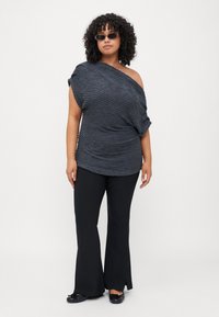 Schwarzes Off-Shoulder-Top aus strukturiertem Stoff, kombiniert mit schwarzen Schlaghosen und schwarzen Ballerinas. Accessoires umfassen rechteckige Sonnenbrillen.