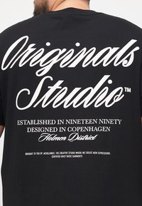 Schwarzes Baumwoll-T-Shirt mit großen weißen Textgrafiken auf der Rückseite. Wichtige Phrasen beinhalten "Originals Studio" und darunter Informationen zum Design.