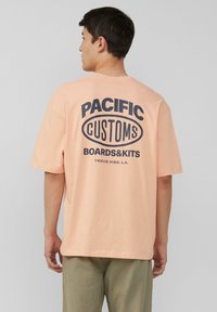 Licht perzik oversize t-shirt met een marineblauwe grafische print op de achterkant met daarop "PACIFIC CUSTOMS BOARDS&KITS VENICE PIER, L.A."