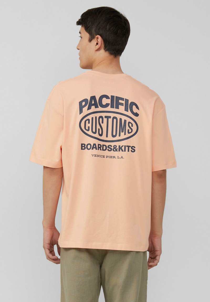 Licht perzik oversize t-shirt met een marineblauwe grafische print op de achterkant met daarop "PACIFIC CUSTOMS BOARDS&KITS VENICE PIER, L.A."
