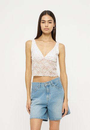 Joven mujer con cabello largo y oscuro que lleva un top corto sin mangas con estampado floral y shorts vaqueros azul claro, de pie frente a un fondo liso.