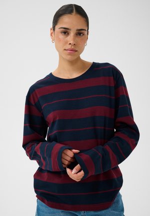 Jeune femme aux cheveux foncés portant un t-shirt à manches longues rayé bleu marine et rouge foncé et un jean bleu, debout devant un fond uni.