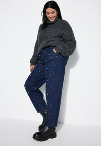 Dunkelgrauer, gemütlicher Strickpullover mit hohem Kragen, kombiniert mit lässigen, dunkelblauen Jeans mit lockerem Schnitt und weißen Perlenverzierungen. Schwarze klobige Stiefel.