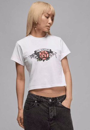 Femme aux cheveux blonds portant de grandes boucles d'oreilles créoles, un T-shirt blanc cropped Air Jordan avec un motif de rose, un jean noir, et une main dans la poche.