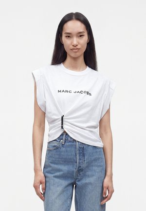 Femme aux longs cheveux foncés portant un t-shirt blanc Marc Jacobs noué à la taille et un jean bleu, debout devant un fond uni.