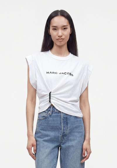 Femme aux longs cheveux foncés portant un t-shirt blanc Marc Jacobs noué à la taille et un jean bleu, debout devant un fond uni.
