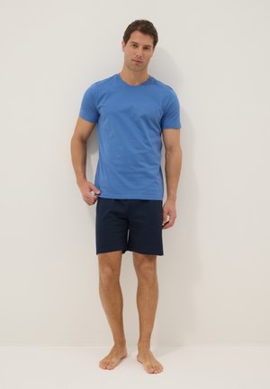 Uomo in piedi a piedi nudi contro un muro bianco, indossa una maglietta blu e pantaloncini blu scuro, con la mano destra che tira leggermente l'orlo della maglietta.