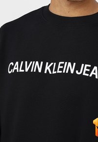 Felpa nera con un evidente logo bianco "CALVIN KLEIN JEANS" sul petto. Scollo tondo a costine, tessuto strutturato.