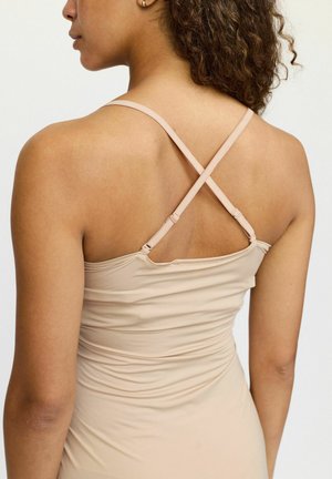 Top sin mangas nude con tirantes entrecruzados, fabricado en una tela suave. Presenta un diseño ajustado y detalles fruncidos en la parte posterior.