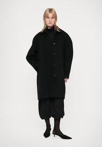 Blond személy, aki túlságosan nagy fekete kabátot, fekete cropped nadrágot, áttetsző fekete harisnyát és fekete hegyes kiscicacipőt visel, fehér háttér előtt áll.