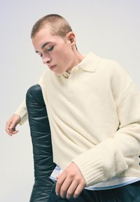 Kremno pulover s pletenico in polo ovratnikom, z tremi gumbi in rebrastimi manšetami. Obučen je čez svetlo modro s karo vzorcem.