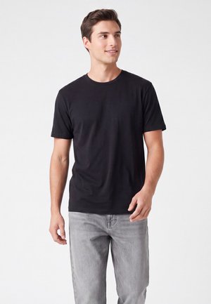 Giovane uomo che indossa una t-shirt nera tinta unita a maniche corte e jeans grigio chiaro, in piedi davanti a uno sfondo bianco uniforme.