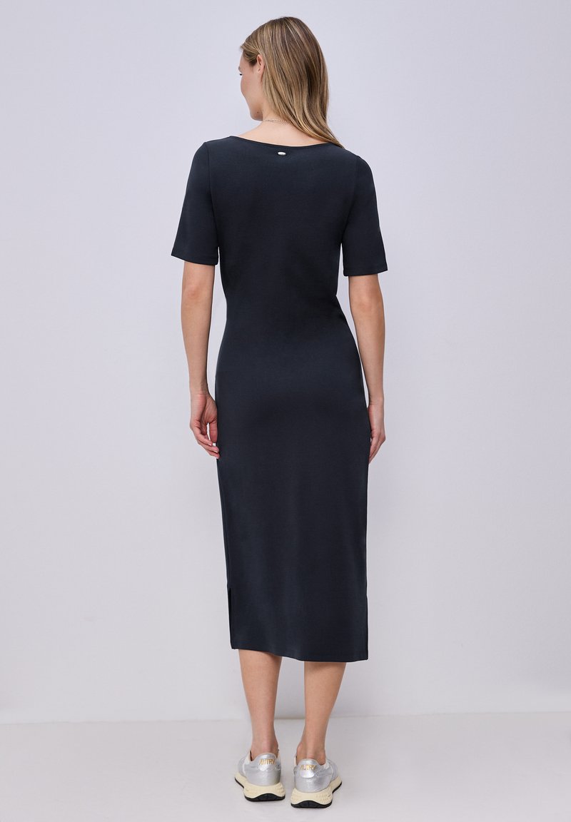 Vrouw staand met de rug naar de kijker, gekleed in een aansluitende zwarte midi-jurk met korte mouwen en zilveren sneakers tegen een effen lichte achtergrond.