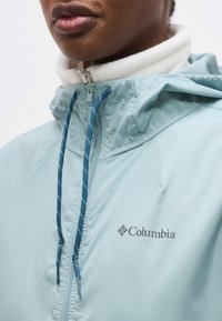 Κοντινό πλάνο ενός ατόμου που φοράει μπουφάν Columbia ανοιχτού μπλε χρώματος με λευκή επένδυση από φλίς και μπλε κορδόνια.