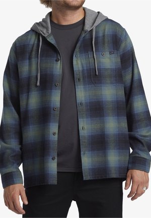 Mężczyzna w niebiesko-zielonej koszuli w kratę zapinanej na guziki z szarym kapturem na czarnej koszulce i czarnych spodniach, ręce rozluźnione po bokach.