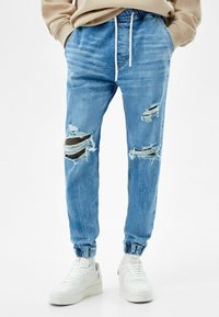 Pantalones jogger de mezclilla de un azul claro con acentos desgastados, puños elásticos y cinturilla ajustable con cordón. Combinados con zapatillas blancas.