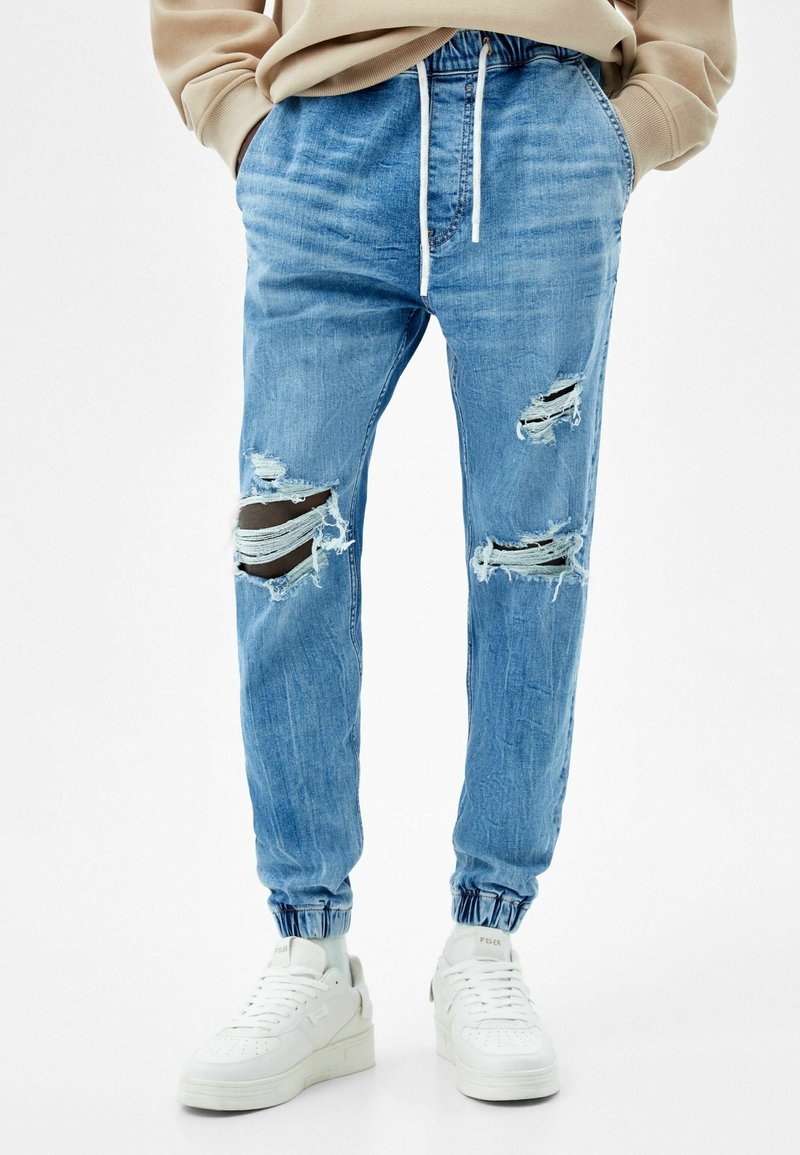 Pantalones jogger de mezclilla de un azul claro con acentos desgastados, puños elásticos y cinturilla ajustable con cordón. Combinados con zapatillas blancas.