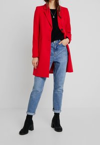 Roter maßgeschneiderter Mantel mit einem einzelnen Knopfverschluss, getragen über einem schwarzen Oberteil und hellblauen Jeans mit aufgerolltem Bund, kombiniert mit schwarzen, hohen Stiefeletten.