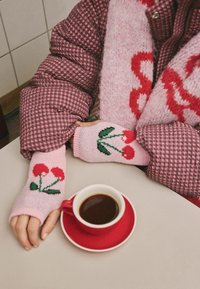 Rožinės megztos pirštinės be pirštų su raudonų vyšnių raštu, derinamos su languota rožine pūkine striuke, šalia raudono kavos puodelio ant stalo.